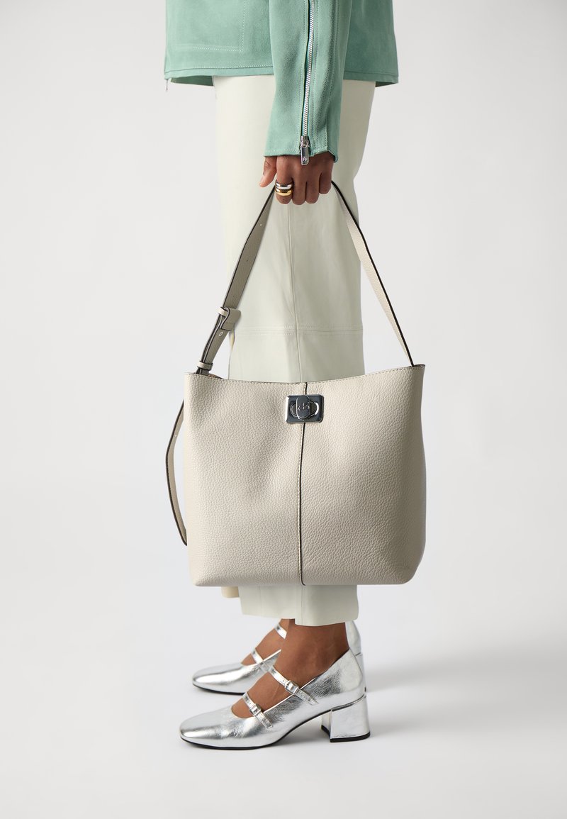 Duża beżowa skórzana torba typu tote o zarysowanej konstrukcji, z zapięciem z srebrnymi elementami i długim regulowanym paskiem. Widoczne srebrne buty na obcasie.