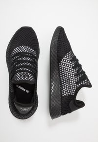 Svarta sneakers i mesh med en texturerad ovandel, utrustade med snörning, en diskret logga och en halvt genomskinlig sula. Modern atletisk stil.