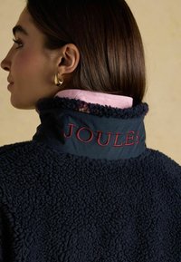 Marineblauwe fleecejas met gestructureerde stof, een hoge kraag met 'JOULES' in rood geborduurd, en een roze gewatteerde voering aan de binnenkant.