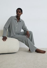 Woman with short hair wearing black and white patterned long-sleeve shirt and matching pants, sitting barefoot on a textured beige ottoman.