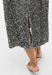 Vestido gris con lentejuelas y diseño de abertura, que presenta lentejuelas circulares brillantes sobre una tela texturizada, y sandalias de tacón metálico.
