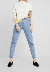 Suéter a rayas en crema y marrón con un acento amarillo, combinado con jeans de denim azul claro y mocasines de ante negros, de pie sobre un fondo gris.