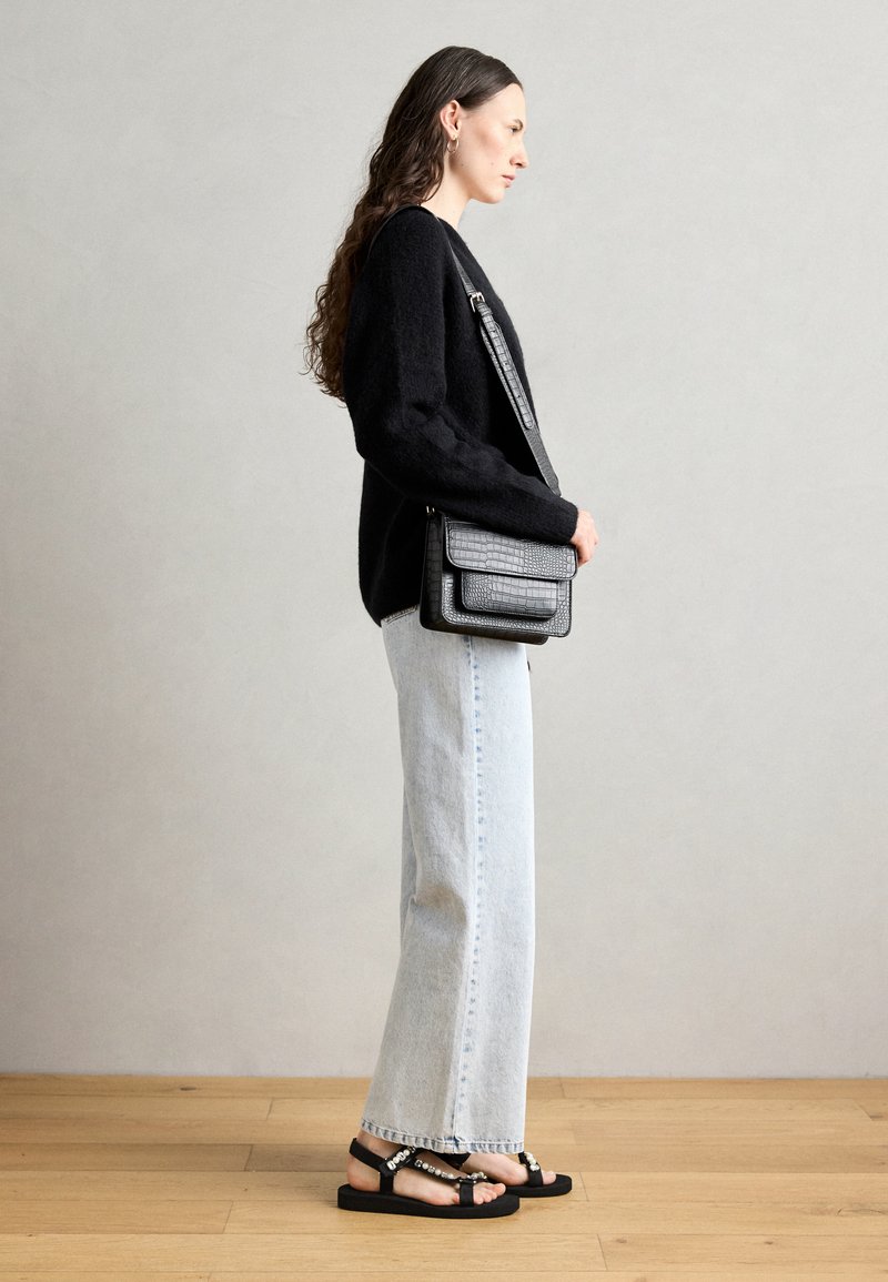 Pull noir texturé avec un col rond, jean bleu clair à jambes larges et sandales noires avec des accessoires décoratifs. Sac bandoulière noir.