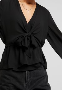 Černá halenka s dlouhými rukávy a hlubokým výstřihem do V, s detailem uzlu vpředu a volánkem na spodním lemu. Hladká, lehká látka.