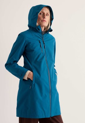Femme portant une veste imperméable à capuche bleu canard, les mains dans les poches, debout devant un fond uni et clair, regardant légèrement vers le haut.