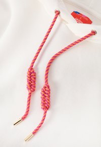 Sudadera blanca con cordones trenzados en rosa y naranja, con aglets metálicos y una etiqueta tejida en el cuello.