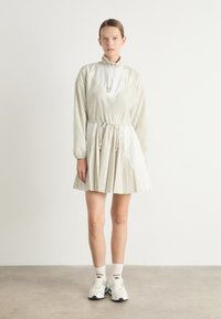 Ljusbeige klänning med hög krage, dragsko i midjan och plisserad kjol, med vita detaljer och långa ärmar. Bärs med sneakers.