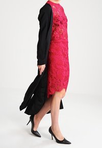 Abito rosso in pizzo con motivi floreali e una giacca nera a maniche lunghe. Il modello indossa tacchi alti neri, mettendo in mostra il design a strati dell'outfit.