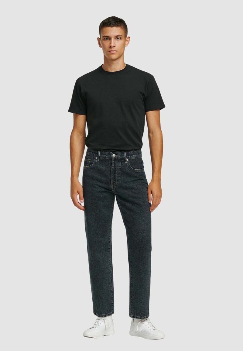 T-shirt nera in cotone con scollo rotondo, abbinata a jeans grigi scuri con tasche frontali. Il modello indossa sneakers bianche. Sfondo neutro.