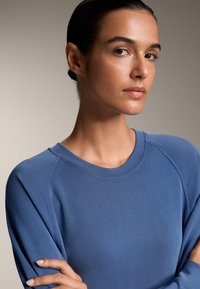 Modra dolga majica s kapuco z mehkimi vlakni, okrožnim izrezom in raglan rokavi. Tkanina se nežno ovija okoli ramen in rok.