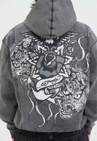 Vista posteriore di una persona che indossa un hoodie grigio con una grafica in stile tatuaggio in bianco e nero di fiori, draghi e il testo "Ed Hardy" sulla schiena.