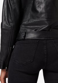 Schwarze Lederjacke mit tailliertem Design, mit Reißverschlüssen an den Ärmeln und einem Gurt in der Taille. Kombiniert mit dunklen Jeans aus Denim mit fünf Taschen.