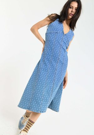 Woman wearing a sleeveless blue polka dot midi dress paired with striped socks and light blue sneakers, standing against a white background.