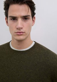 Maglione a coste verde oliva con scollo rotondo, indossato sopra una t-shirt grigia chiara. Texture liscia e design aderente.