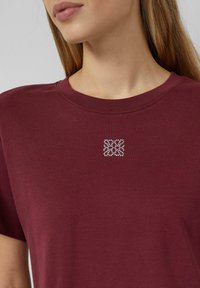 Bordo kokvilnas t-krekls ar apaļu izgriezumu un īsām piedurknēm, ar nelielu kristālu rotātu ģeometrisku dizainu uz krūšu zonas.