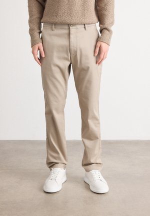 En person klädd i beige byxor, vita sneakers och en strukturerad beige tröja som står på ett slätt grått golv mot en vit vägg.