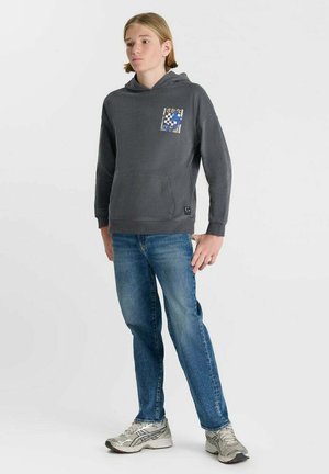 Jonge persoon die een donkergrijze hoodie met een afbeelding op de borst draagt, blauwe spijkerbroek en witte sneakers, staand tegen een effen witte achtergrond.