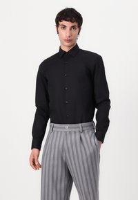 Black button-up shirt with long sleeves, smooth texture, paired with gray vertically striped pleated trousers. Notable button details.