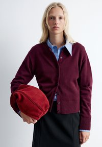 Bordeauxfarbener Cardigan mit Knopfleiste und gerippten Manschetten, über einem hellblauen Hemd getragen. Das Model hält eine rote Wildledertasche vor einem schlichten Hintergrund.