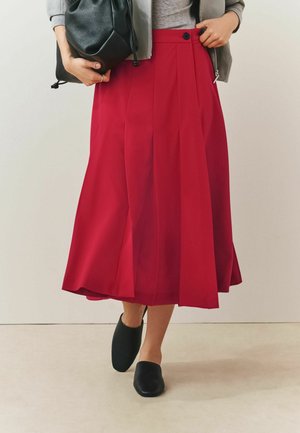 Frau trägt einen roten plissierten Midirock, graues Oberteil, graue Jacke, schwarze Slipper und hält eine schwarze Ledertasche.