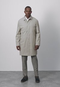 Abrigo impermeable beige claro con cuello de solapa, dos bolsillos delanteros y cierres ocultos, usado sobre pantalones grises y zapatos oscuros.