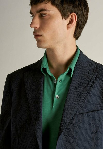 Blazer blu navy in tessuto con un motivo distintivo, sovrapposto a una polo verde con tre bottoni. Linee pulite e vestibilità su misura.