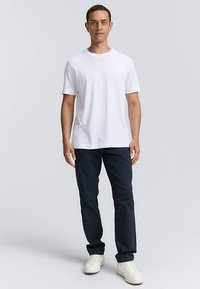 White cotton t-shirt paired with dark navy trousers, worn with cream-colored sneakers. Model stands with arms relaxed at sides.