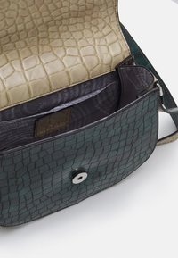 Zielona skórzana torba o teksturze, z wzorem krokodyla, beżowym klapą, zapięciem na zatrzask oraz ciemną kieszenią wewnętrzną z wzorem. Regulowany pasek na ramię.