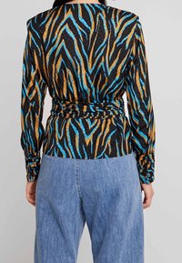 Top a maniche lunghe nero con motivo a zigzag blu e oro. Presenta un design avvolgente, tessuto testurizzato e maniche arricciate con bottoni decorativi.