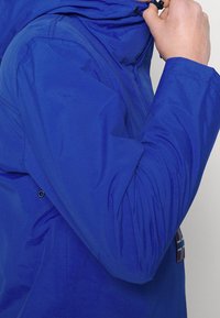 Osoba upravující kapuci jasně modré bundy s viditelným švem a malou nášivkou poblíž pasu.