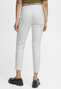 Modèle féminine de dos, vêtue de pantalons ajustés gris clair et de chaussures noires à enfiler, portant un haut décontracté vert olive, se tenant devant un fond blanc.