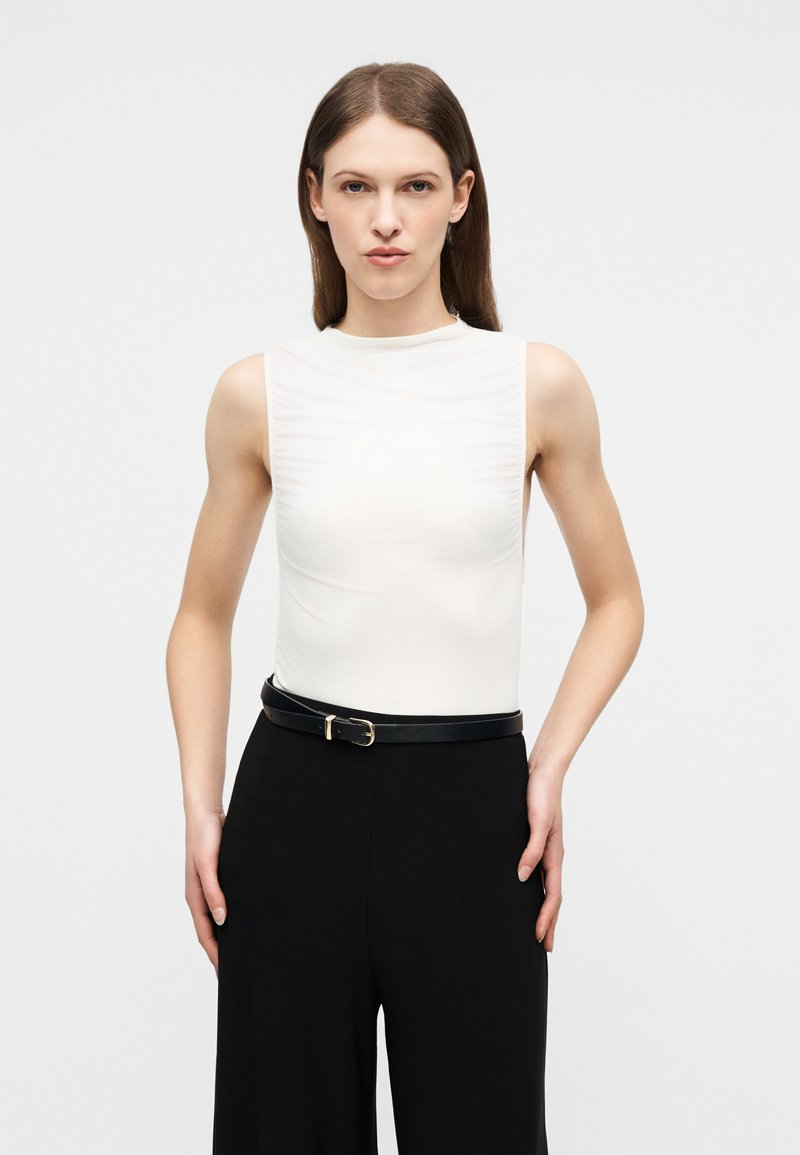 Femme aux cheveux bruns lisses portant un haut blanc sans manches rentré dans un pantalon noir avec une ceinture noire, debout devant un fond uni.