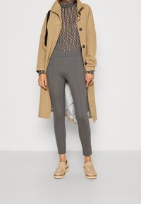 Beige trenchcoat över svart och vit randig polotröja, grå leggings och beige loafers med träsålar, med slät textur och åtsittande design.