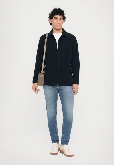 C�ămașă button-up de culoare albastru închis, blugi de culoare albastru deschis, adidași bej cu tălpi maro. Modelul ține o poșetă mică de umăr de culoare bej. Design și texturi simple.