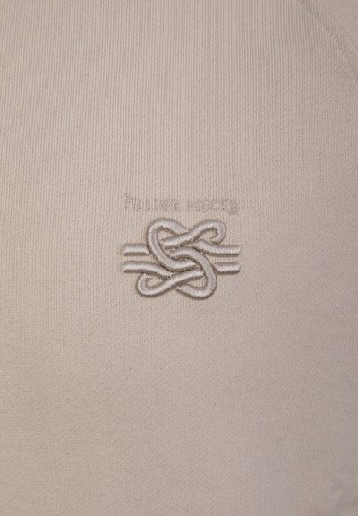 Béžová látka s vyšívaným abstraktním uzlovým vzorem a slabým nápisem nad ním.