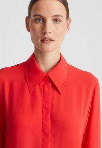 Rotes Shirt mit großem, spitzem Kragen und vorderer Knopfleiste; aus einem glatten, leichten Material mit einfacher Textur gefertigt.