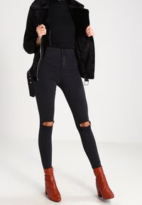 Svarta högmidjade jeans med slitna detaljer vid knäna, tillsammans med en svart ribbad topp och en svart jacka med konstig pälsfoder. Bruna klackiga ankelstövlar.