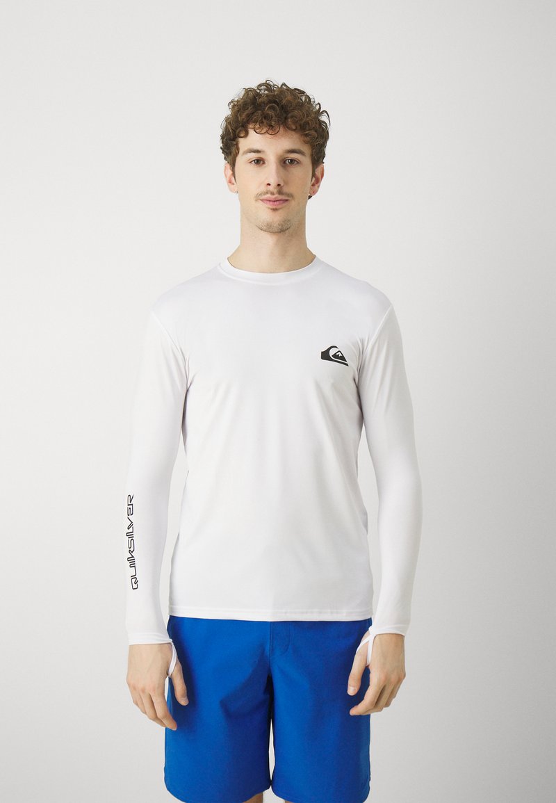 Balts garām piedurkņu rasšsargu ar melnu logotipu uz krūtīm un "Quiksilver" uzrakstu uz piedurknes, kombinējot to ar ziliem šortiem. Strukturēta, stiepļu auduma.