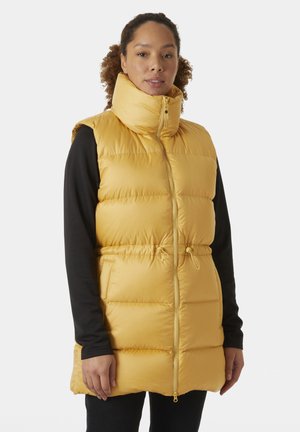 Chaleco acolchado amarillo con cuello alto, cierre de cremallera y cintura con cordón ajustable. Presenta una textura suave y bolsillos laterales, superpuesto sobre una camiseta negra.