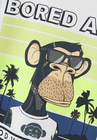 Hvid t-shirt med et grafisk design af en abe med solbriller. Baggrunden viser gradientfarver i grøn og blå med palmetræer.