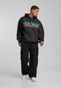 Black hoodie with colorful dripping "Zoo York" graphic, paired with black cargo pants. White sneakers complete the outfit. Simple, urban style.