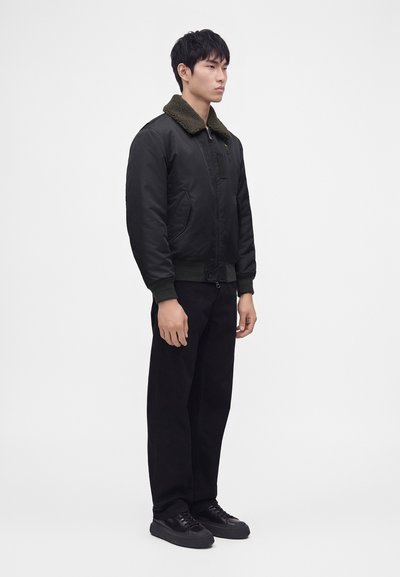 Jeune homme debout face à droite, portant une veste bomber noire avec un col en fourrure foncé, un pantalon noir et des baskets noires sur fond blanc.