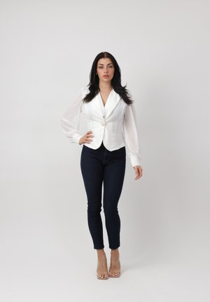 Femme debout portant un blazer blanc ajusté avec des manches transparentes bouffantes, un jean skinny foncé et des chaussures à talons hauts transparentes sur un fond uni.