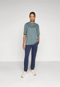 Kvinne med flette som har på seg en løs turkis t-skjorte, marineblå joggebukser og hvite sneakers, står med den ene hånden i lommen.