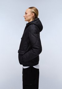 Svart puffjacka med huva, med en quiltad struktur, framfickor och elastisk nederkant. Modellen bär svarta byxor och en vit skjorta.