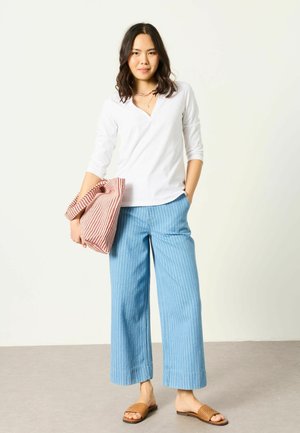 Frau steht, trägt ein weißes langärmeliges Oberteil, helle, weit geschnittene Hose mit Streifen, braune gewebte Sandalen und hält eine rot-weiß gestreifte gefaltete Tasche.