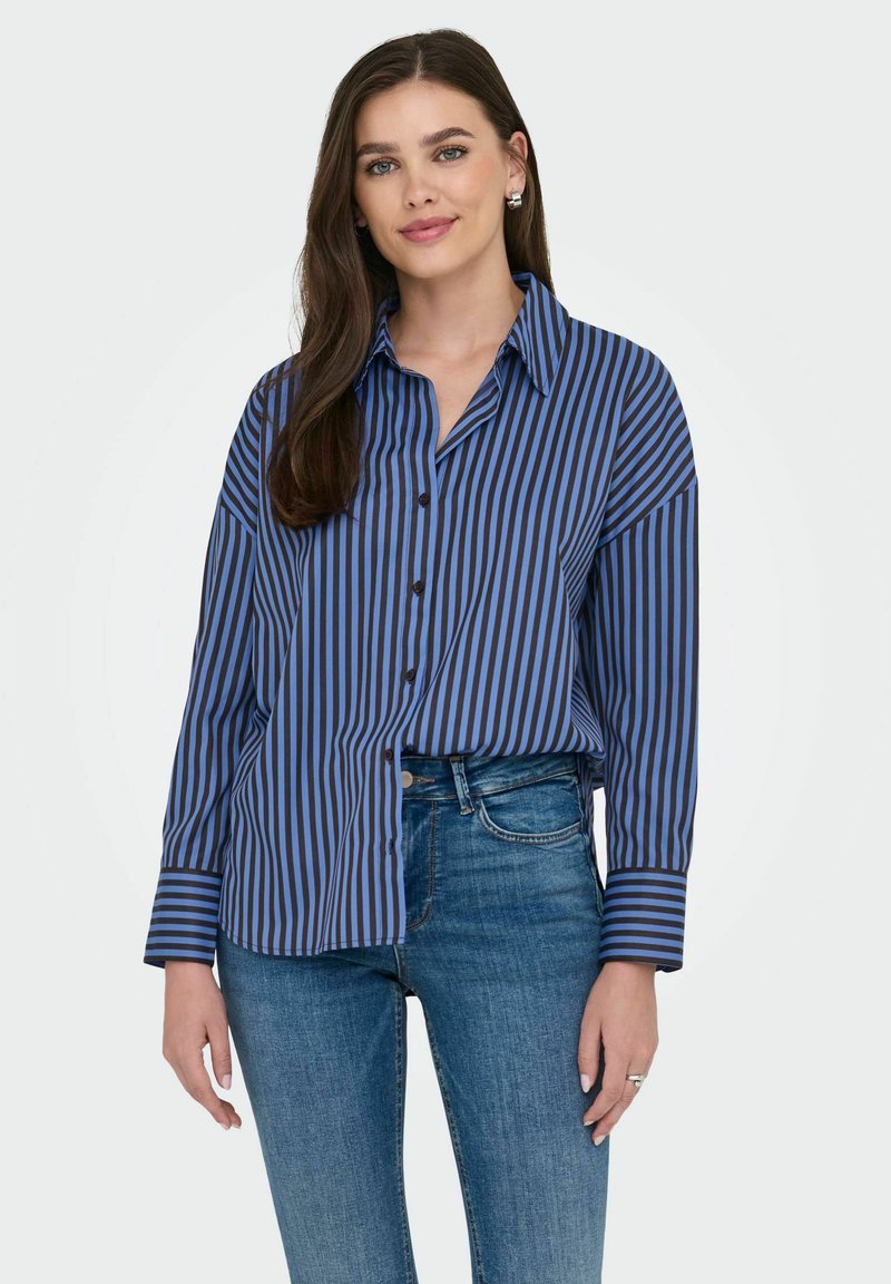 Chemise à rayures bleues et noires, à boutons, avec col et manches longues, présentant une coupe décontractée et une texture lisse, portée avec un jean bleu.