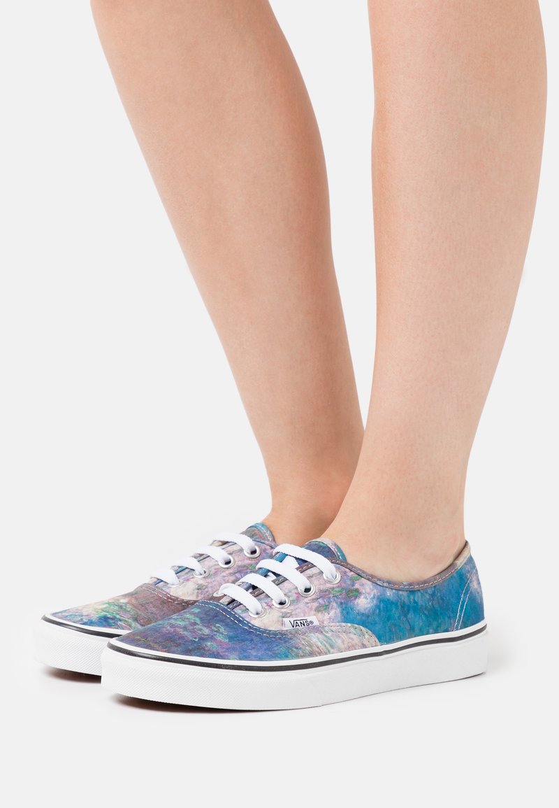 Pieds portant des baskets Vans basses avec un motif tie-dye multicolore bleu-violet et des lacets blancs sur un fond blanc.