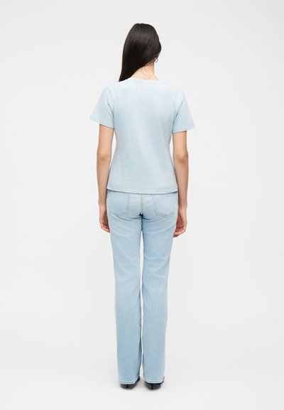 Γυναίκα με μακριά σκούρα μαλλιά, που φοράει ανοιχτό μπλε κοντομάνικη μπλούζα και ανοιχτό μπλε τζιν παντελόνι με ίσια μπατζάκια, στέκεται πλάτη σε μονόχρωμο φόντο.