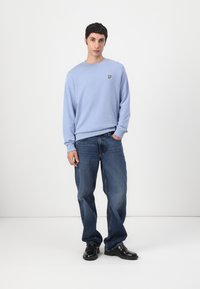 Sudadera de algodón azul claro con cuello redondo y un logo de mariposa amarilla. Combinada con vaqueros azules de corte holgado y zapatos negros.